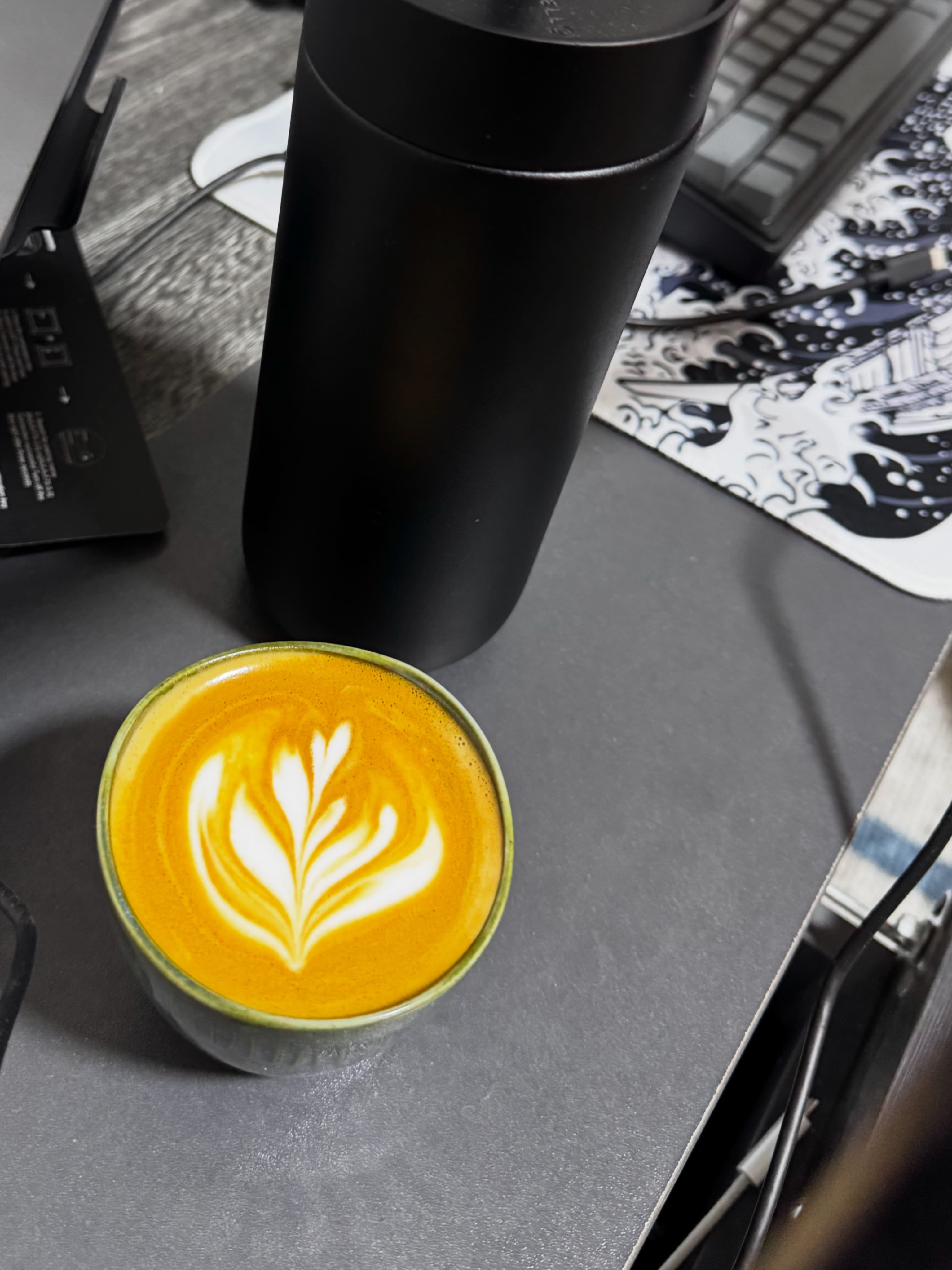 A cup of coffee with latte art and a black flask on a desk.